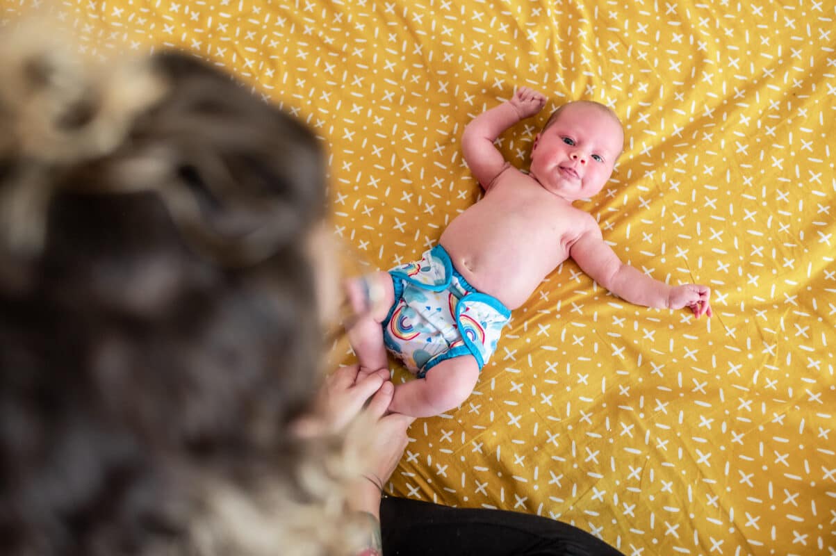 Baby mit blau-weißer Stoffwindel-Überhose mit Regenbogenmuster auf senfgelber Wickelunterlage.